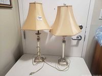 Pair of two table lamps on a white table in front of a door, showing overall lamps with beige fabric shades and metallic bases.