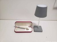 Pen in red-lined case next to small gray desk lamp with cylindrical shade