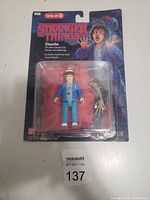 Photo of the Stranger Things Dustin action figure in original packaging with an accessory arm piece. Packaging is intact but partly damaged.