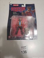 Photo of the Lucas action figure from Stranger Things in its original packaging that shows significant wear and tear, partially torn at edges, with figure and accessories visible inside.