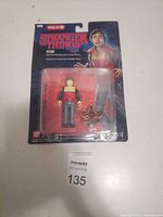 Front view of Stranger Things Will action figure in original plastic and cardboard packaging showing figure and accessory.