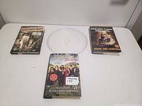 All items on white table: three Duck Dynasty and Duane Dog Chapman books and a clear round microwave glass plate in center.