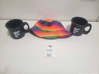 Two black speckled mugs with white F logos alongside a vibrant rainbow bucket hat on a white table.