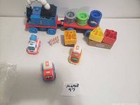 Photo showing Thomas the Tank Engine blue train with red cargo car loaded with barrels and several small character toy vehicles and accessories arranged on white surface.