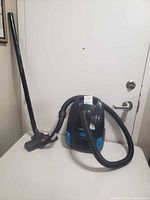 Front view of a black and blue canister vacuum cleaner with a flexible hose, wand, and floor brush attachment placed against a white door.