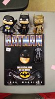 Photo showing three Batman Funko Pop vinyl figures and a Batman hardcover book on a gray surface.