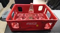 Red plastic Coca-Cola crate holding a single clear glass Coca-Cola bottle with original labeling and cap.