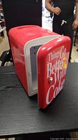 Front and side view of the red Coca-Cola mini fridge showing door with 'Things Go Better With Coke' text open, revealing the interior space. The small fridge sits on a table with a person's partial figure in the background.