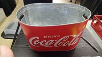 Photo of a red, rectangular Coca-Cola metal drink tub with handles on each side, featuring the white Coca-Cola logo and "Drink Coca-Cola" text.