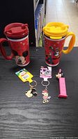 Two Disney-themed travel mugs, two keychains, and one Minnie Mouse PEZ dispenser arranged on a black table surface.