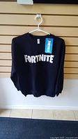 Front view of black Fortnite sweater hanging on hanger with white Fortnite logo visible and original tag attached.