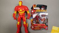 Photo shows two Iron Man figures: a large red and yellow articulated Iron Man figure and a smaller figure in original packaging labeled 'Iron Man 2', also predominantly red and yellow.