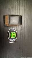 Photo showing a brown leather case and a metal John Deere belt buckle with green and yellow logo placed on a wooden surface.