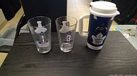 Photo showing two Toronto Maple Leafs themed clear glasses and one insulated travel mug on a dark table surface.