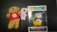Photo showing the Disney Pop! vinyl figure in box alongside two plush toys, yellow bear with red 'h.' shirt and pink bird plush.