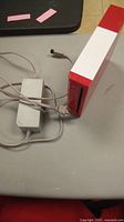 Red and white Nintendo Wii console with power cord on a flat surface. View shows console standing vertically with power cord connected.