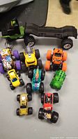 Photo of nine toy monster trucks including one large green and black truck with flatbed trailer and eight smaller trucks in various colors and designs.