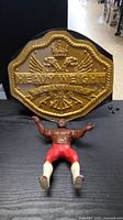 Photo showing a gold-colored wrestling heavyweight championship belt with embossed eagle and crown design, and a wrestling figure in red shorts and white boots lying on a black surface.
