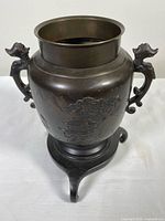 Front and side view of Japanese bronze urn showing raised relief landscape and architectural motifs, two sculpted handles, and tripod pedestal