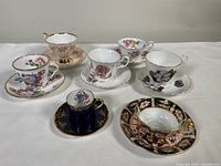Seven different antique porcelain teacups and saucers together on a white cloth background, showing different floral patterns and colors.