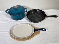 Shows the blue enamel round Dutch oven with lid, black cast iron skillet, and white oval enamel skillet on wooden tray together.