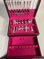 Open wooden chest showing flatware set arranged inside with upper rack holding knives and bottom compartment holding various spoons, forks, and serving utensils.