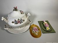 White ceramic tureen with lid and handles decorated with ceramic flowers, displayed with matching oval under-plate and three rectangular ceramic wall plaques with floral designs mounted on fabric backgrounds.