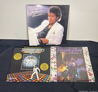 Three vinyl record albums arranged on black background: Michael Jackson Thriller, Bee Gees Saturday Night Fever, and Prince Purple Rain.