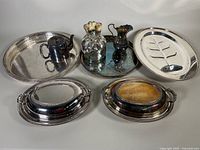 Overview of various silver plated servingware items arranged on a light background including platters, covered dishes, teapot, and decorative containers.