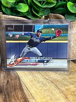 Front of 2018 Topps Ronald Acuna Jr rookie card showing him fielding and Rookie Debut date, in protective case.