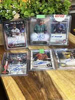Six autographed baseball cards displayed on wood surface with green plants in background