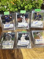 Six Bowman Topps Chrome baseball cards with autographs, displayed in clear plastic holders with green tape at the top, against a green plant background.