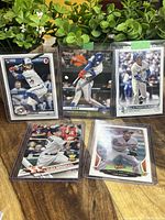 Photo showing all five rookie cards displayed on a wooden surface with green plants in the background. Cards are in protective sleeves.