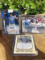 Three signed baseball cards displayed in protective cases, placed on a wooden surface with greenery in the background.