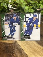 Two rookie hockey cards standing side by side, one of Mitch Marner and one of William Nylander, on a wooden surface with a green leafy plant in the background.