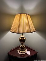 Brass table lamp turned on, showing warm light through a pleated beige fabric lampshade on a polished brass base, sitting on a wooden table with crochet doily.