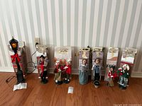 Full set of 7 Christmas caroler figurines standing in front of original boxes alongside the plug-in lamp post with red bow and greenery decoration.