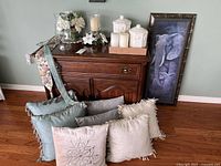 Wide view showing framed elephant art print, wooden cabinet, six throw cushions in front, white ceramic canisters on cabinet, glass candle holder with artificial floral decor.