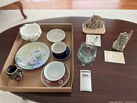 Photo of mixed lot showing various teacups and saucers, small decorative sculptures, hand-painted plate, and glass bowl with spoon on a wood table.