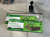 Photo showing two boxed Urban Gardener expandable metal raised garden beds, an outdoor lantern, and two metal shepherd hooks lying on the ground.