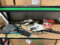 Photo showing 2 pairs of white ladies size 7 ice skates, multiple tennis racquets stacked on a shelf, and Oakley goggles.