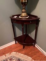 Dark wooden corner accent table positioned in a corner, showing the overall structure, top, drawer, and lower shelf with a lamp on top.