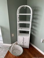 Tall white wicker shelf with four open shelves and two-door cabinet below, shown with white wicker basket in front.