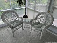 Two white wicker armchairs flanking a round wicker side table with glass top in a sunroom