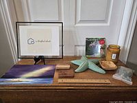 Original signed art framed with house sketch and text, ceramic starfish, wooden bowl, coasters, candle, vase filler, wooden ruler, and garden calendar displayed on a wooden surface against a door.