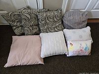 Eight assorted pillows arranged on floor including swirl patterned, pink plain, white patterned, Disney princess themed, and buttoned gray pillows.