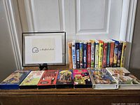 Photo of assorted VHS movie cases arranged next to photo display with 'a thankful studio' text, showing movies including Toy Story and others laid flat in front.