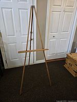 Wooden collapsible easel shown fully extended standing on floor next to a door. Visible wear and paint marks on the wood surfaces.