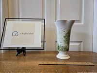 Tall white ceramic vase with a flared opening and handpainted green trees and grass, black birds flying, shown next to ruler for scale.