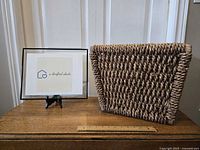 Front view of the large woven basket placed on a wooden surface beside a framed logo with a ruler for scale.